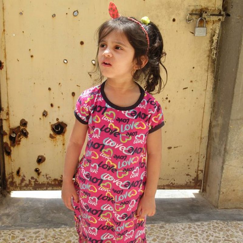 A little girl, wearing a dress with the word ‘love’ printed on it in a repeating pattern, stands in front of a bullet-hole riddled door.