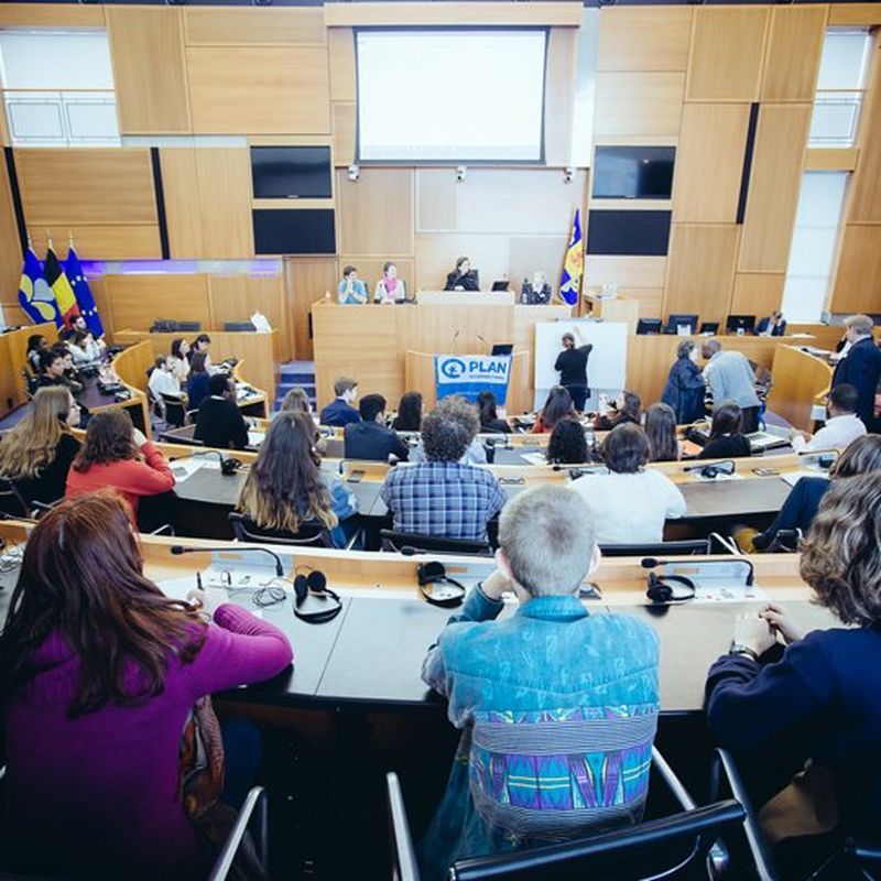 Three rows of people sitting with their backs to the camera in a semi-circle, facing a podium and a Plan International banner.