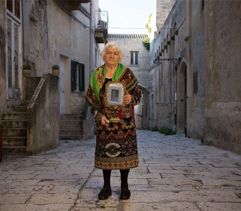An older, shawled woman stands on a cobbled street, holding a photograph in a frame.