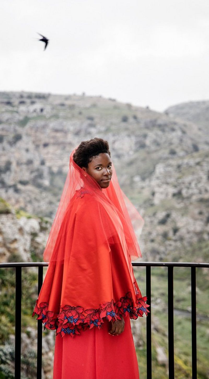 A woman in a red dress, shawl and veil, stands by railings, overlooking mountains.