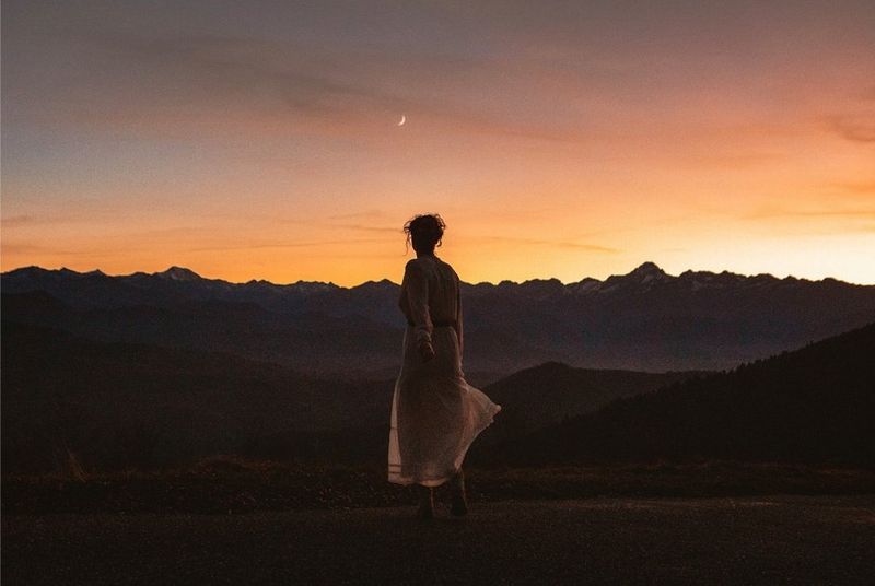 A woman stands with her back to the camera, watching an orange sunset over dark mountains. She wears a pale dress, and her hair is worn up, with tendrils outlined in shadow.