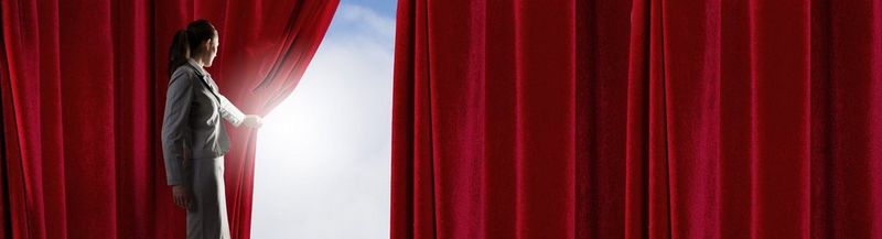 A woman wearing a business suit pulls back a red curtain to reveal a bright blue sky and clouds.