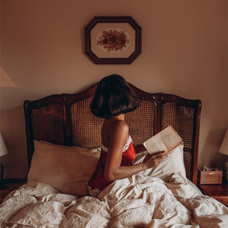 A woman sits up in bed, with her back to the camera. She has shoulder length brown hair in a bob and wears a red satin camisole with white lace trim and holds what appears to be a notebook. Above the dark wood vintage rattan headboard is are pressed flowers in an octagonal dark wood frame.