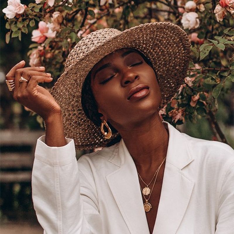 A woman in a white suit and wide-brimmed straw hat sits in front of a blossoming tree. Her right hand is raised, and her eyes are closed. She wears two gold rings, two gold necklaces and gold drop earrings.