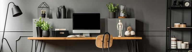 A stylish grey-walled home office, with a light wood desk and chair, several plants, a lamp, objects on black shelves and a desktop computer.