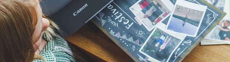 A photograph from above. A blonde child wearing a green and white checked school dress stands next to a table, upon which a Canon printer is printing out a scrapbook page of photos with the title ‘festival’. 