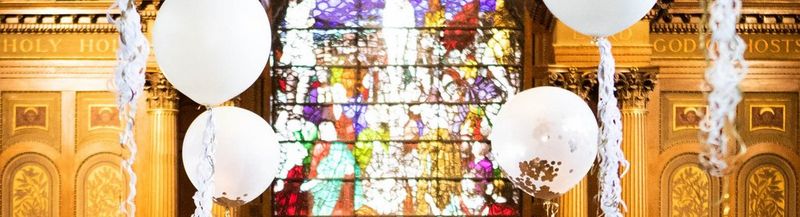 Balloons attached to streamers float in front of ornate golden walls and a complex, multicoloured stained glass window.
