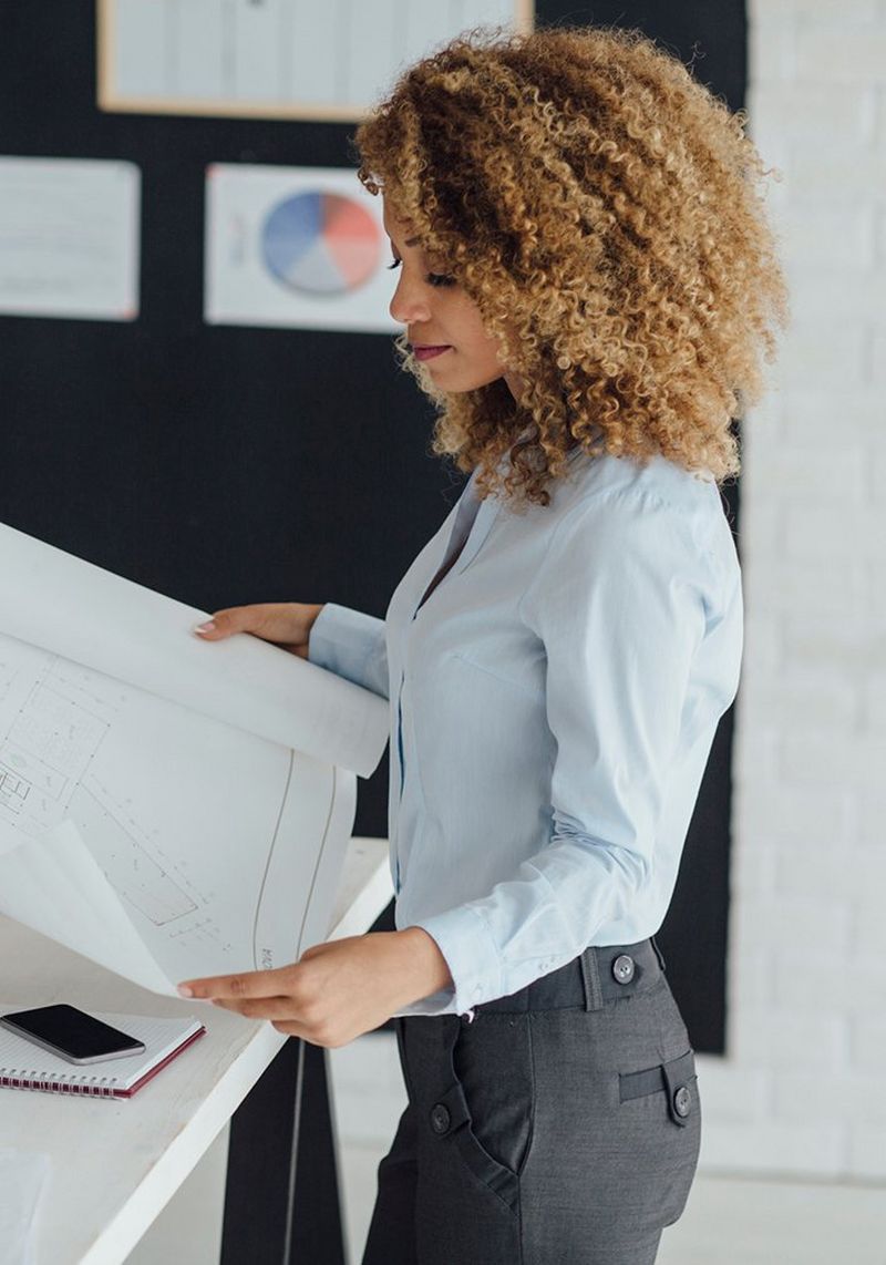 Woman with curly hair, wearing a light blue shirt and dark grey trousers, stands at a white desk with laptop, mug and pens, looking at a large piece of paper.