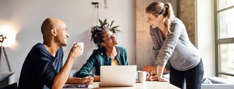 Woman in grey jumper leans hands on table, chatting with a bald man and a woman wearing a green cardigan who sit in front of a laptop, tablet device and phone. 