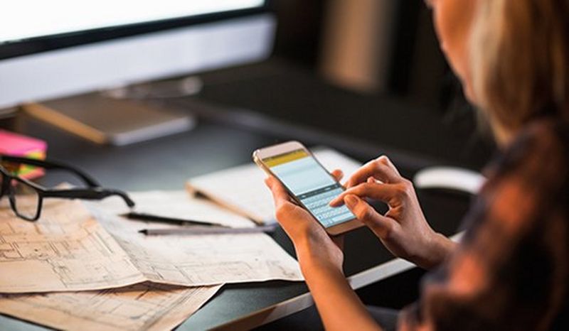 Out of focus woman sits at a black desk with desktop computer, papers and a pair of black glasses, typing on a smartphone. 