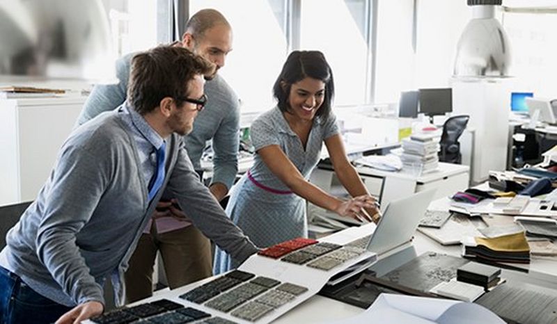 Two men and a woman wearing business smart/casual look at textile swatches and a laptop on a long desk covered in books, papers and folders. 