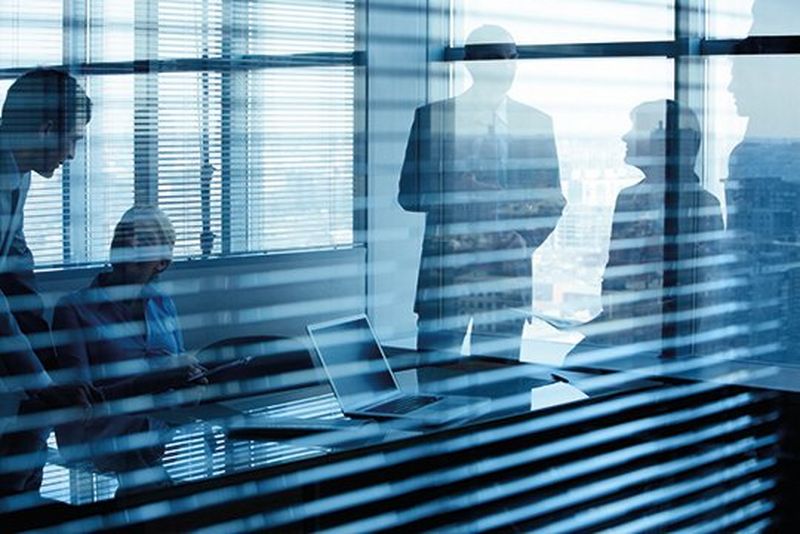 A blue-tinged view through a window with slatted blinds into a meeting room with the shadow/outlines of four people standing and sitting around a table with a laptop.