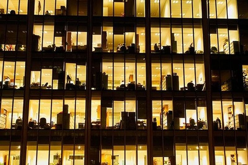 A view into an office building at night, with the windows lit up in yellow and office equipment in silhouette.