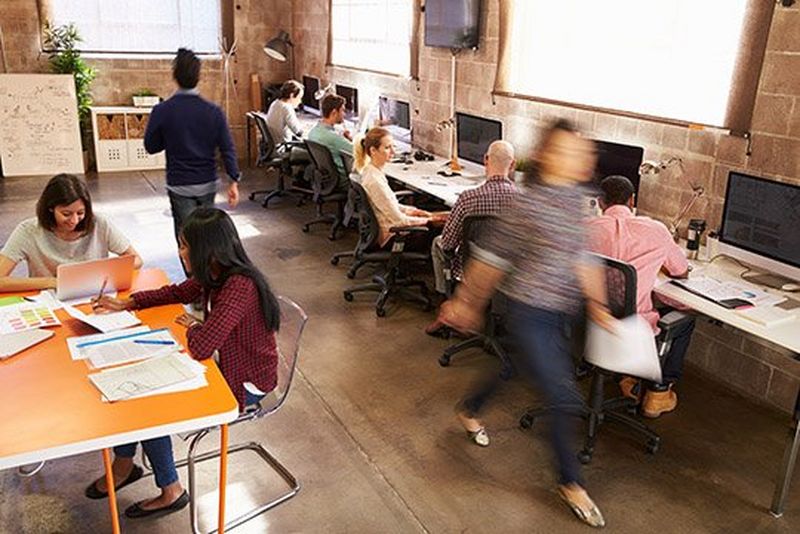 A casual office environment with people working at their computer desks or a communal table, and a blurry woman rushing past.