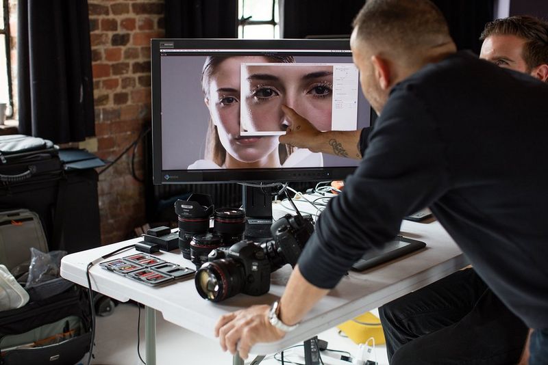 A man leans over a desk and points at an image of a woman's face on a monitor. A Canon camera and lenses are also on the desk. 