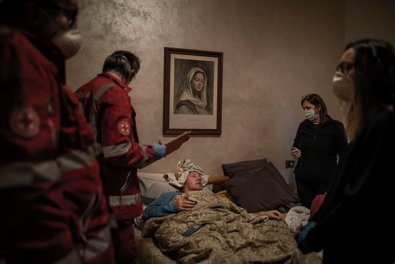A man lies in bed surrounded by Italian Red Cross volunteers. A painting of the Virgin Mary hangs on the wall. Photo by Fabio Bucciarelli.