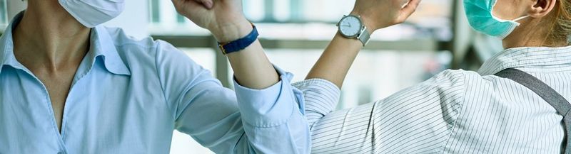 Two women elbow bump in greeting. Their faces are not clearly seen, but they are both wearing face coverings, work attire and watches.
