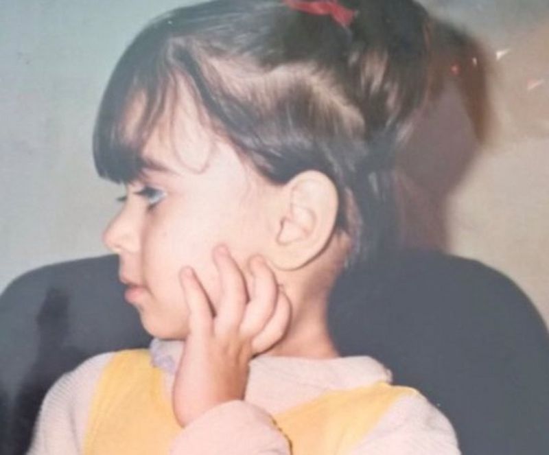 A head and shoulders photo of a little child, wearing a white and yellow top and with a high ponytail, held with a red bow. She is facing left and has one hand raised to her cheek.