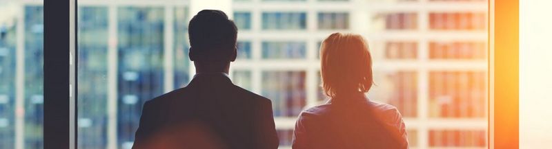 A man and woman, dressed in office attire and standing with their backs to the camera, looking out of a window at other office buildings.