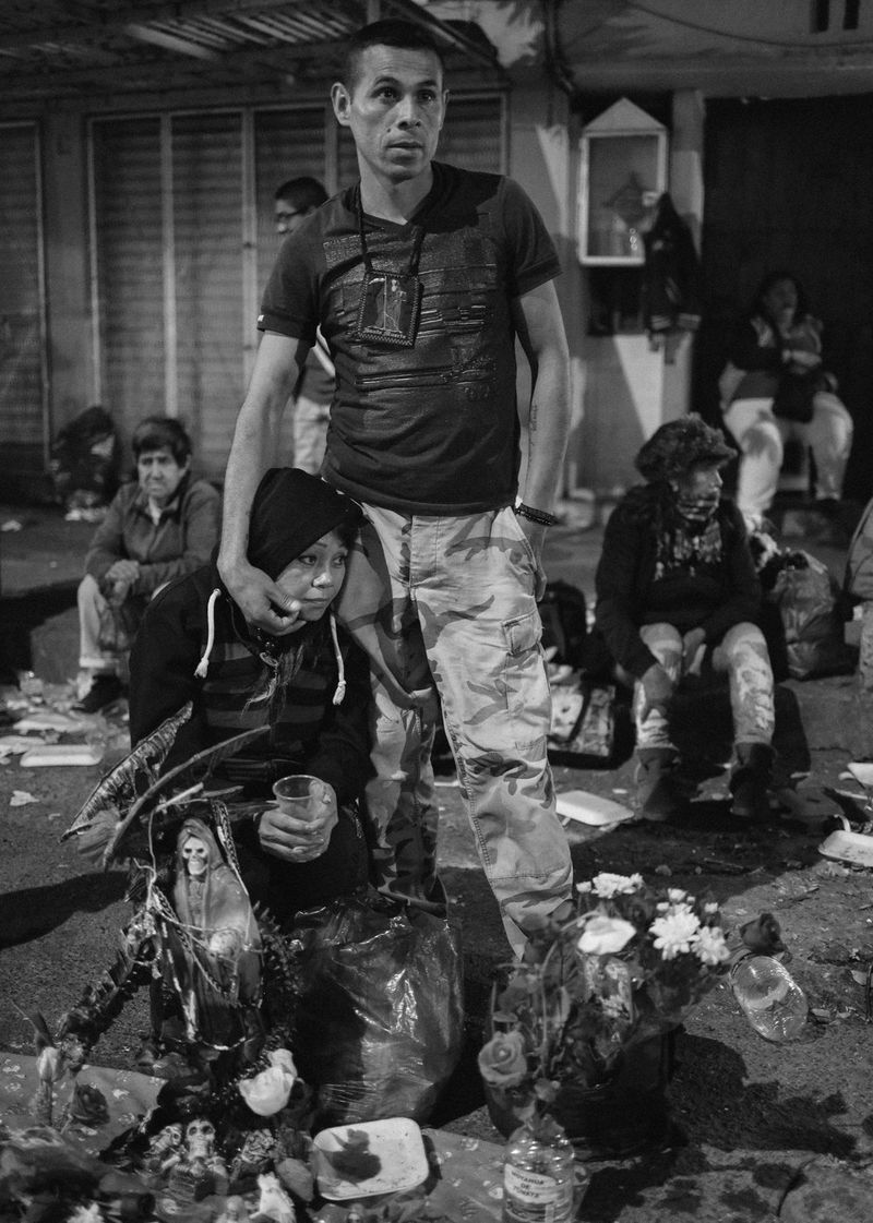 A man stands next to a kneeling woman, his hand gently on her cheek. They are surrounded by statuettes of Death, flowers and various offerings.