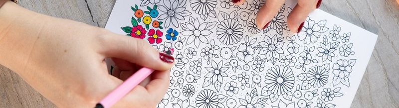 A pair of hands with short, dark red painted nails rest on a partially filled colouring sheet of black and white illustrated flowers.