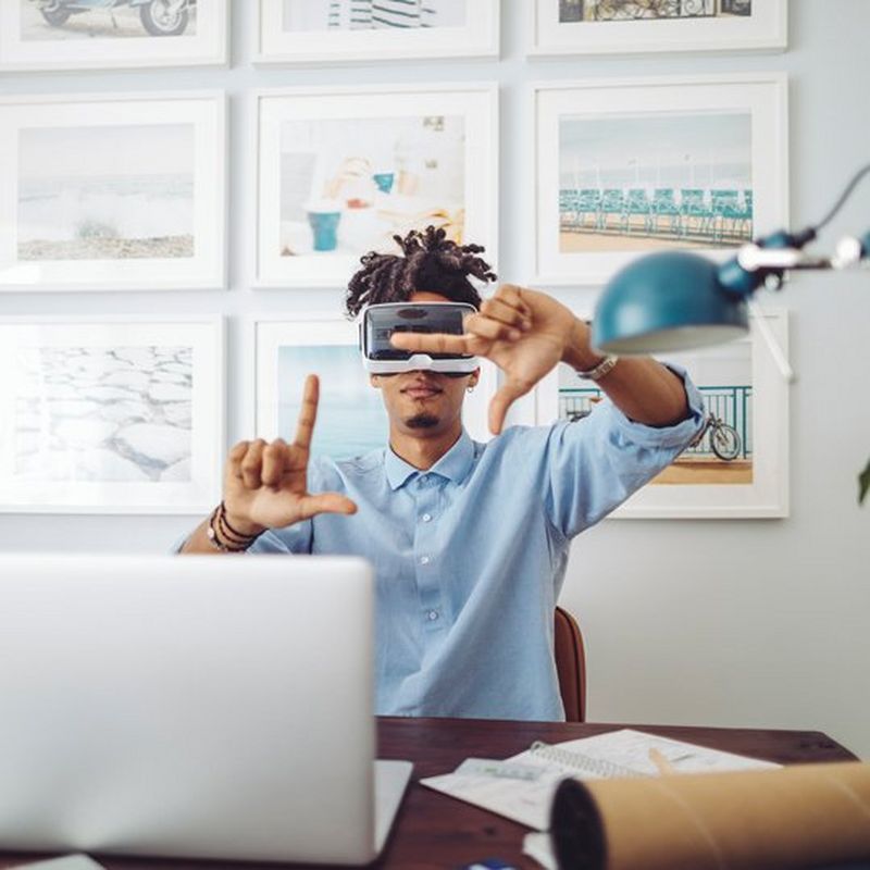 A young man sits at a desk wearing a VR headset and holding his fingers in a ‘frame’ pose. In front of him is a laptop, notebook and lamp. Behind him are nine equally sized pieces of artwork in white frames.