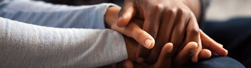 A close up of two people’s hands, on top of each other. They rest on a denim-clad knee. The arms in shot are covered by blue-grey sleeves.