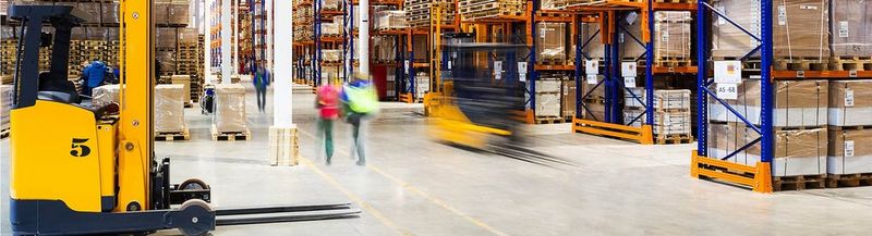 An action shot of a warehouse, with forklift trucks and people moving between huge racks of boxes on pallets, awaiting shipment.