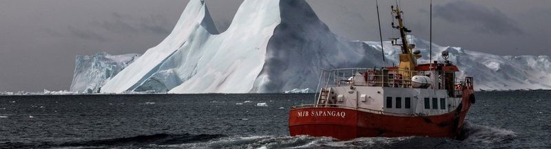A red boat sails on cold grey water, with a huge floating iceberg behind it.