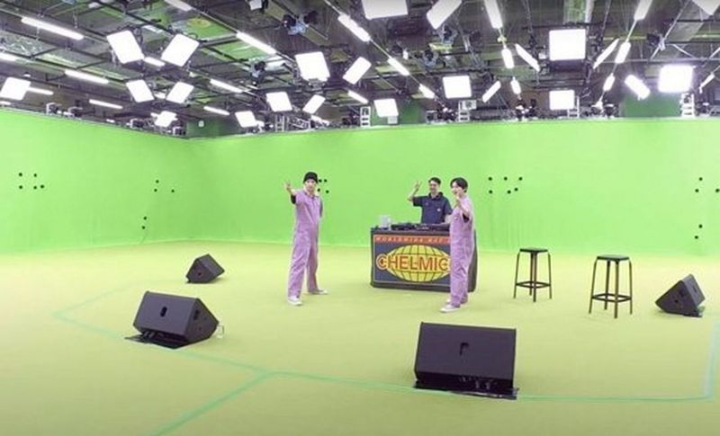 Two women in lilac jumpsuits stand in a green-screened studio. They each have an arm outstretched. Behind them is a man standing behind a DJ booth that is labelled ‘chelmico’. © Warner Music Japan Inc. 