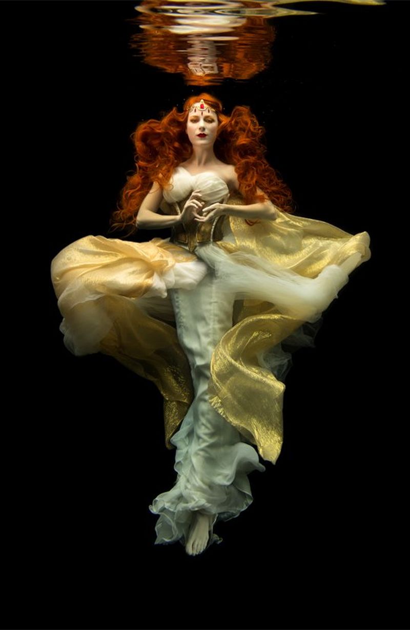 A red-haired woman floats against a black background, wearing a diaphanous dress of gold and pale yellow. 