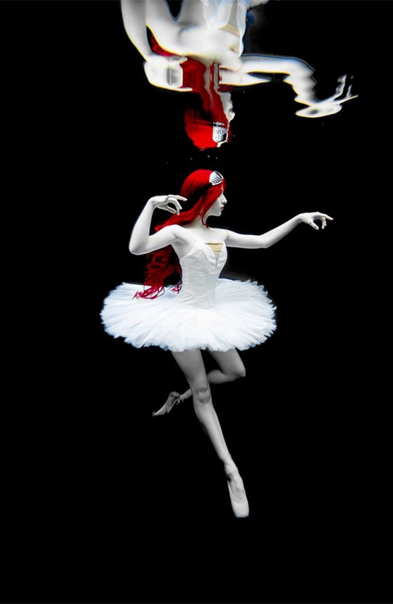 A ballet dancer with pillar-box red hair and alabaster skin, floats en pointe against a black background, wearing a pure white tutu. 