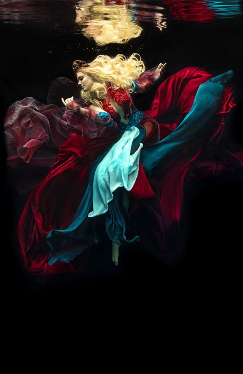 A golden-haired woman in a many-layered gown of blood red and sea blues, floats against a black background. 