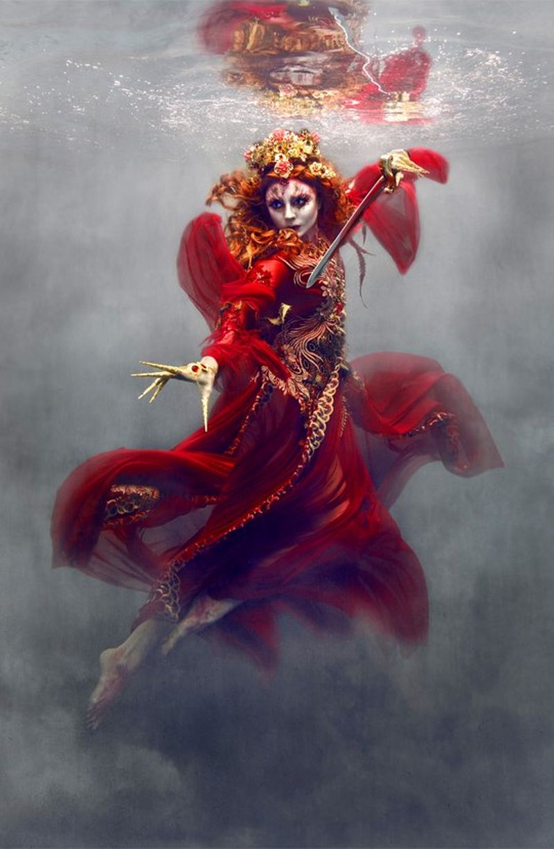 A woman with a painted face floats in clouds and wears an eastern-style red gown with a semi-mandarin collar. Her red hair is decorated with flowers and she assumes the battle position, directing a sword at an unknown opponent to the left-hand side of the image.  