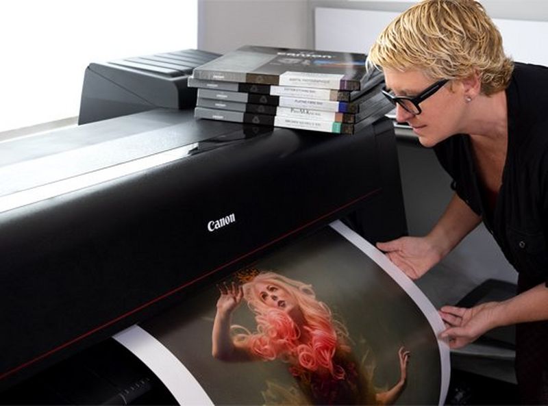 Cheryl stands to the right, with short, blonde hair and black framed glasses. She gently holds a print as it emerges from a Canon PRO-4100 printer.