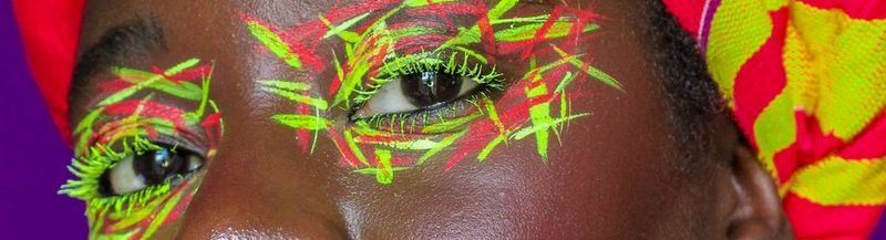 A woman’s eyes, painted with a neon yellow and red crosshatch pattern. She wears a matching head-wrap, which you can see just in shot. © Wendy Asumadu