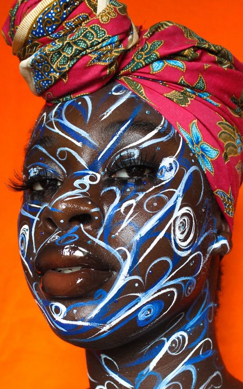 A woman wearing a red head-wrap that is printed with green and blue leaves and flowers. Her face is painted with swirls of blue and white and her lips are high gloss.