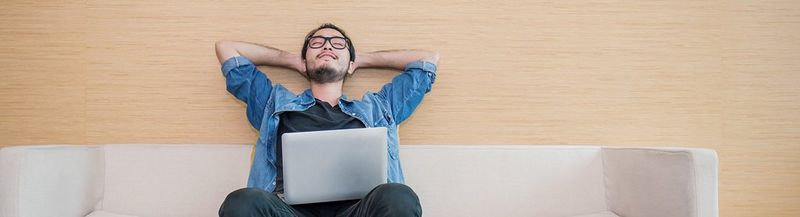 A young man in glasses reclines on a couch with his laptop resting on his lap.
