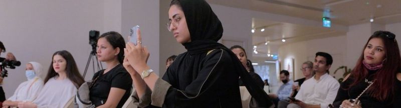 Women and men sitting in an audience. In the centre of the image is a woman dressed in black, wearing glasses and a head covering, using her phone to film what she is watching.  