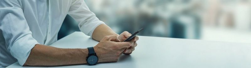 The torso of a man in a white shirt, sat at a white table, holding a mobile phone. He has a black watch on his right wrist.