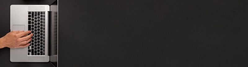 A thin landscape image that is plain black for nearly the entire space, except for the far-left hand side, where a silver laptop can be seen, viewed from above. Its screen can barely be seen, and the keyboard faces the left of the image. On the keyboard sits a hand and wrist, the rest of the arm out of shot.