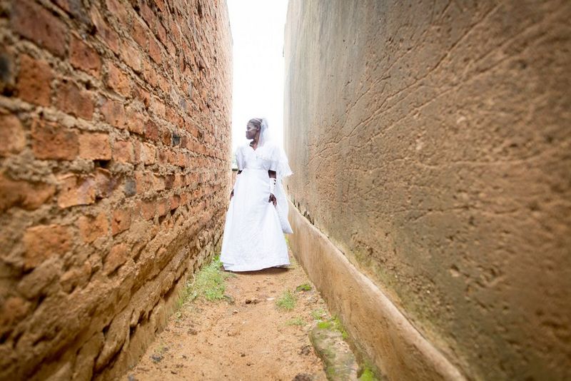 A smiling bride in Kenya, wearing her wedding dress, is photographed in an alley between red brick walls. 
