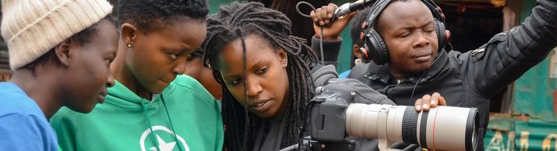 Three young women looking at the rear display of a camera, while a man in headphones behind holds a boom mic.