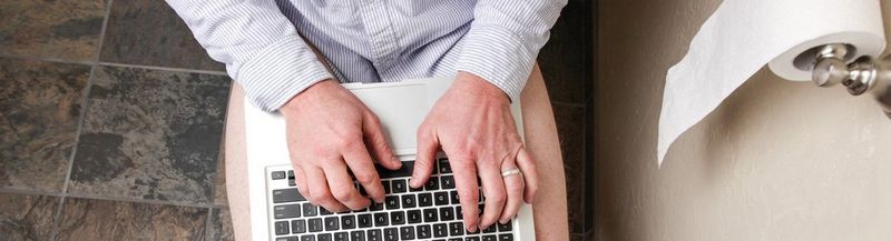 A top-down view of a man in a business shirt, sitting on the toilet. His thighs are bare, but with his laptop on his lap. To his right is a toilet roll on a wall-mounter holder.