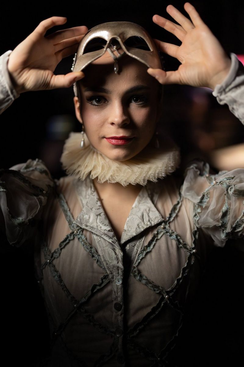 Een persoon in een theatraal kostuum met een kraag met ruches en doorschijnende mouwen met een patroon houdt een zilverkleurig masker boven het gezicht en kijkt rechtstreeks naar de camera.