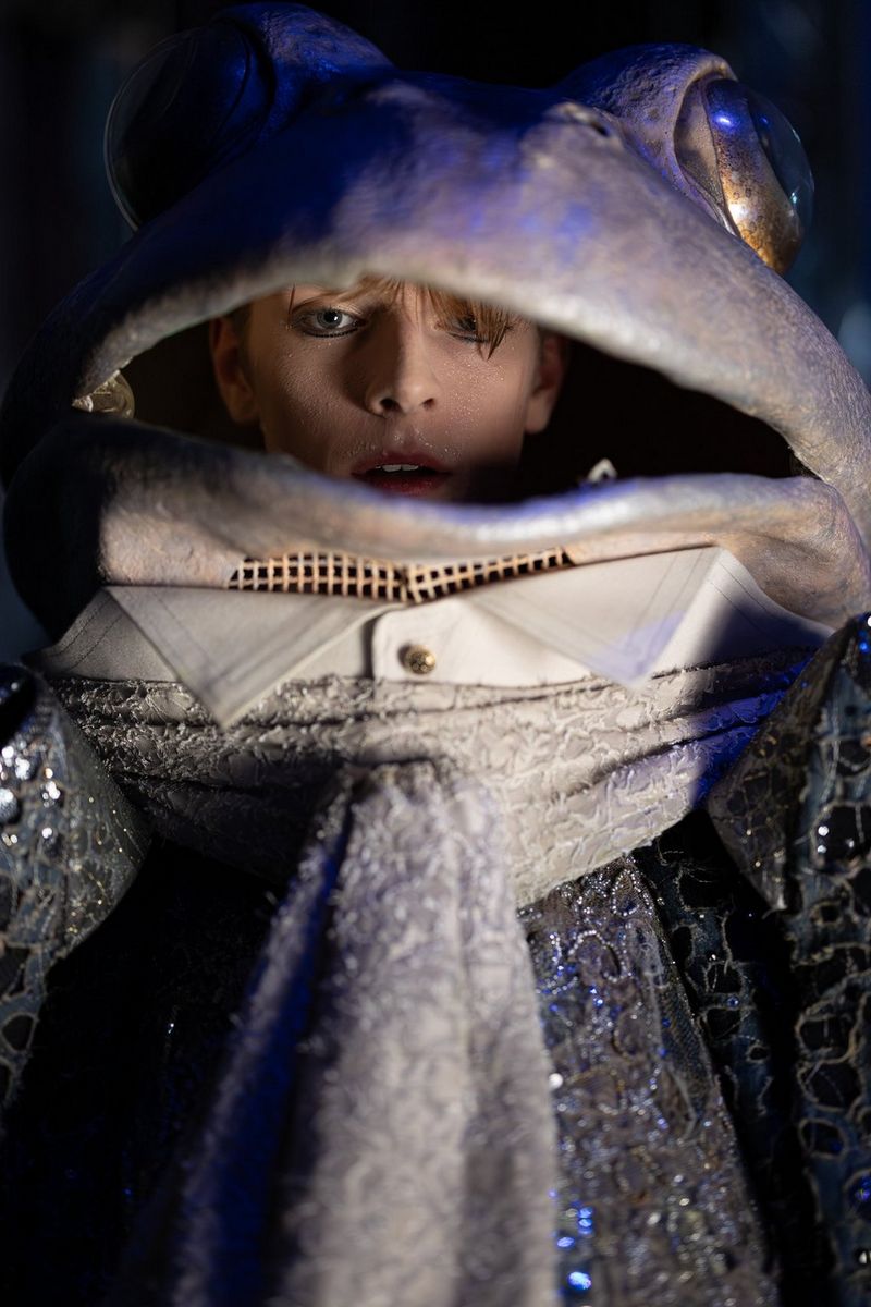 Een close-up vanuit een lage hoek van het gezicht van een danser met veel make-up, die door de open mond van een groot kikkerkostuum met pailletten kijkt.