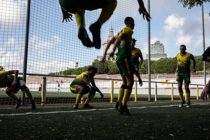 A group of football players in yellow and green strips during a training drill. One player is mid-jump, others are squatting or mid squat Behind them are nets, green trees, and a partly cloudy sky. Courtesy of Goals For Change Project. Created by Ofelia de Pablo, Javier Zurita and Pablo Tosco.