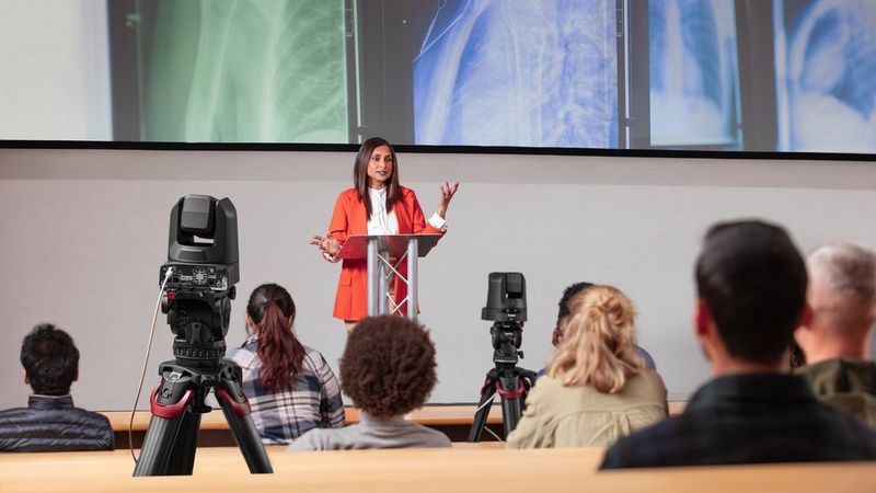 Zwei Canon PTZ-Kameras auf Stativen im Publikum übertragen einen Livestream einer Vortragenden, die vorne steht. Hinter ihr sind farbige Röntgenbilder zu sehen.