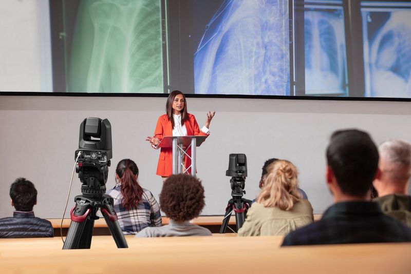 Two Canon PTZ cameras on tripods positioned among the audience members are livestreaming a presenter standing at the front, with coloured X-ray images displayed behind her.