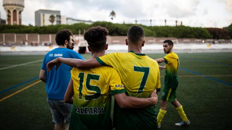 Two football players stand on an artificial turf field, facing away from the camera with their arms around each other's shoulders. Behind them to the left, a person in a blue shirt and light-coloured shorts stands with their back to the camera, while another player in a similar yellow and green uniform is slightly blurred in motion to the right. Courtesy of Goals For Change Project. Created by Ofelia de Pablo, Javier Zurita and Pablo Tosco.